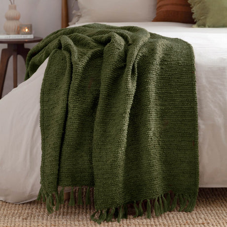 Candar Plain Polycotton Throw in khaki green, boucle-textured with fringed edges, draped over a white bed in a bedroom setting.