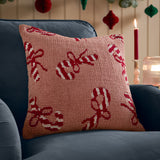 Square tufted cotton cushion in pink with red and cream candy cane and bow pattern, shown on a navy sofa beside a lit candle.