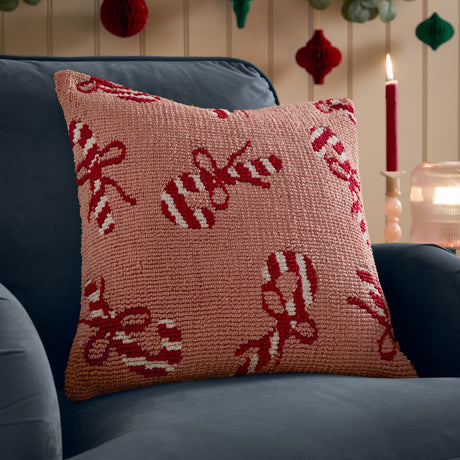 Square tufted cotton cushion in pink with red and cream candy cane and bow pattern, shown on a navy sofa beside a lit candle.