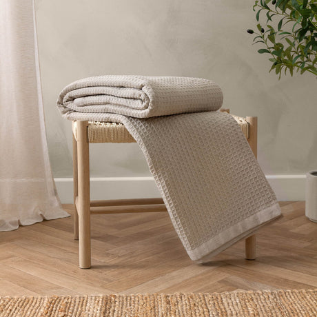 Canopy Plain Cotton Throw in natural, cotton waffle weave with plain border, draped over a wooden stool in a room with woven rug and curtain.