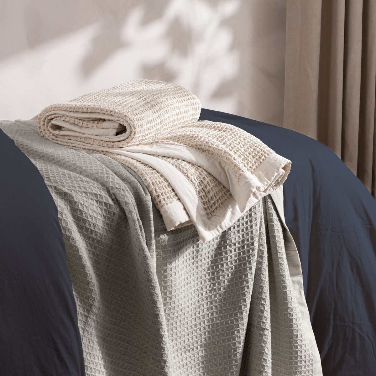 Canopy Plain Cotton Throw in natural, folded chunky waffle-weave cotton with a plain woven border, draped over a grey waffle blanket on a bed with navy linen.