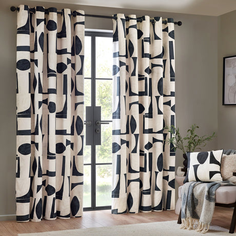 Carro Abstract Lined Eyelet Curtains in Dusk, cream cotton panels with bold black geometric shapes, hung on a black rod beside a glass door in a living room.