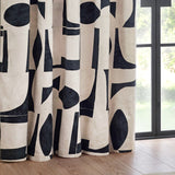 Carro Abstract Lined Eyelet Curtains in Dusk, cream cotton fabric printed with bold black geometric shapes, hung in eyelet folds beside a window.