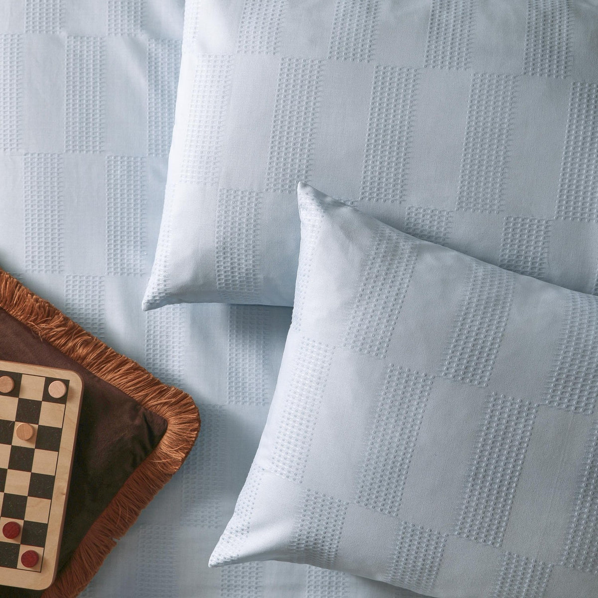 Club Checkerboard duvet cover set in pale Blueberry blue, showing waffle-weave check texture on duvet and pillowcases with a checkers board and pieces nearby.