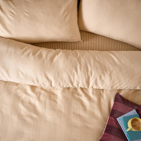Club Checkerboard duvet cover set in latte, showing waffle-textured squares on the duvet and pillowcases with a striped reverse, styled on a bed with a book and mug.