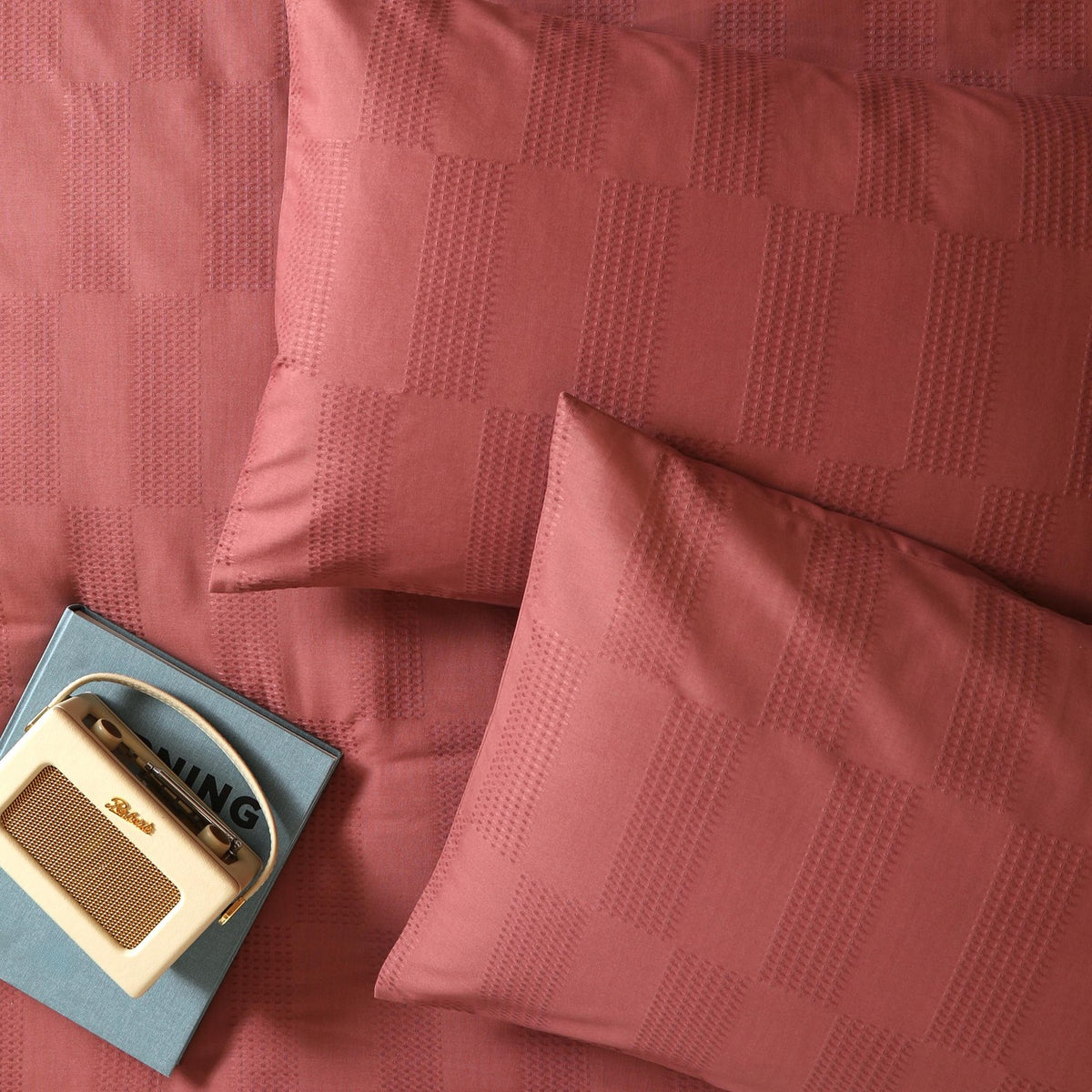 Club Checkerboard duvet cover set in raspberry pink, showing waffle-textured checkerboard weave on duvet and pillowcases, styled on a bed with a vintage radio and book.