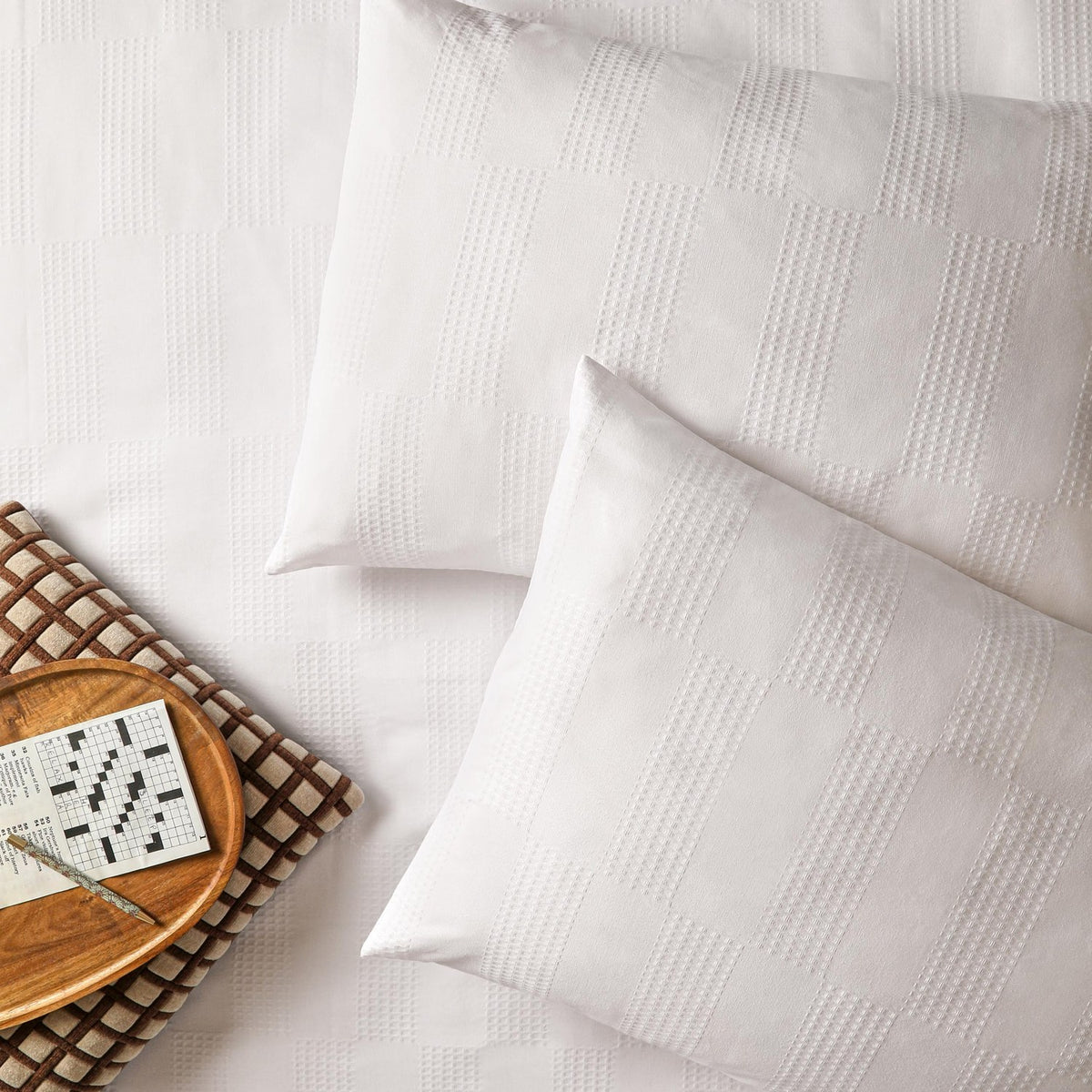 Club Checkerboard duvet cover set in white with waffle-textured checkerboard pattern, shown with matching pillowcases on a bed beside a wooden tray and crossword puzzle.