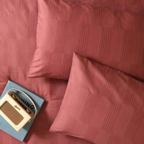 Club Checkerboard cotton blend pillowcase pair in raspberry red with woven waffle-texture check design, styled on a bed with a vintage radio and book.