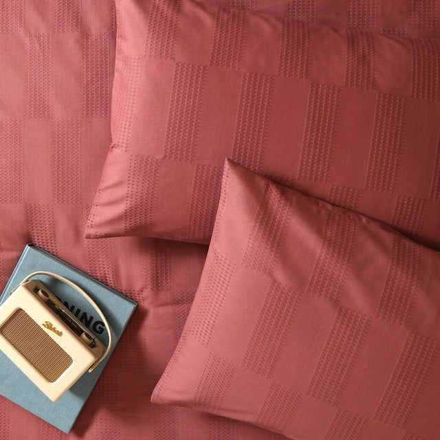Club Checkerboard cotton blend pillowcase pair in raspberry red with woven waffle-texture check design, styled on a bed with a vintage radio and book.