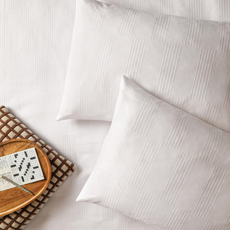 Club Checkerboard cotton blend pillowcase pair in white with a woven waffle check texture, styled on a bed with a wooden tray and crossword puzzle.