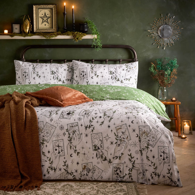 Celestial Stories reversible duvet cover set on a bed, cream side with fine-line tarot cards, hands, moons, stars and green foliage, styled with a green pillowcase and rust cushion.