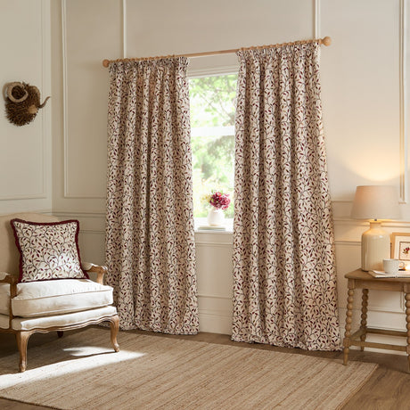 Cervino pencil pleat curtains in cream jacquard fabric with a trailing damson red, purple and green leaf design, shown hanging in a living room beside a matching cushion.