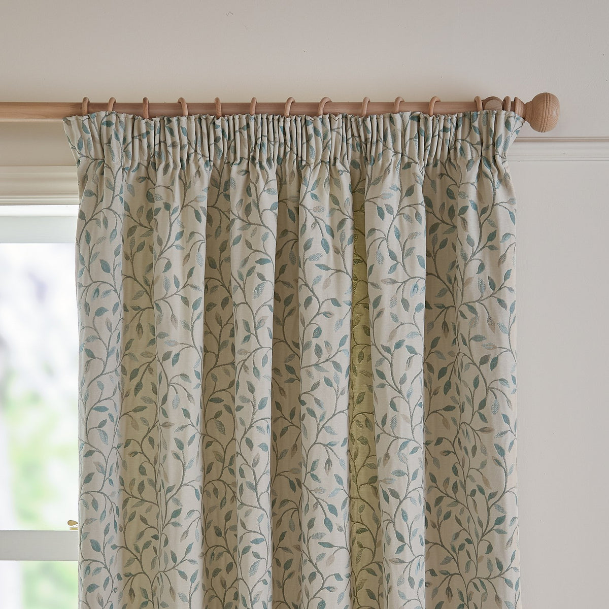 Cervino Jacquard Pencil Pleat Curtains in duck egg, cream fabric with woven trailing leaf pattern in blue, sage and tan, hung on a wooden pole near a window.
