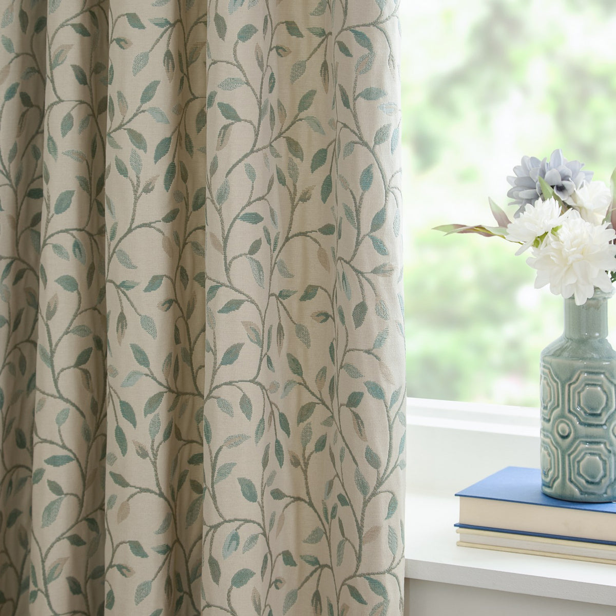 Cervino Jacquard Pencil Pleat Curtains in Duck Egg, cream fabric with woven trailing leaf design in blue, sage and tan, shown pleated by a window beside a vase of flowers.