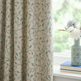 Cervino Jacquard Pencil Pleat Curtains in Duck Egg, cream fabric with woven trailing leaf design in blue, sage and tan, shown pleated by a window beside a vase of flowers.