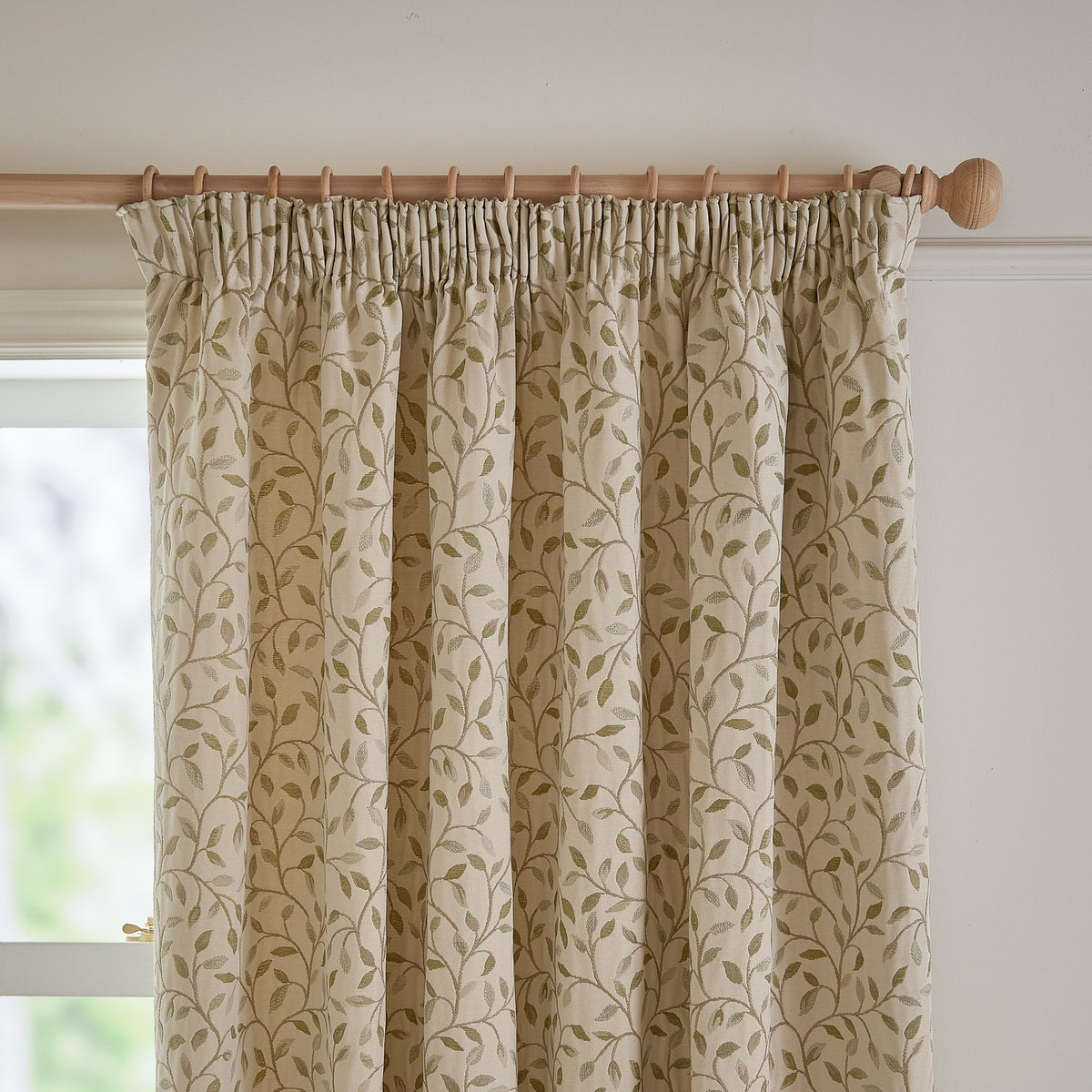 Cervino pencil pleat curtains in cream jacquard fabric with trailing green leaf pattern, shown pleated on a wooden pole beside a window.