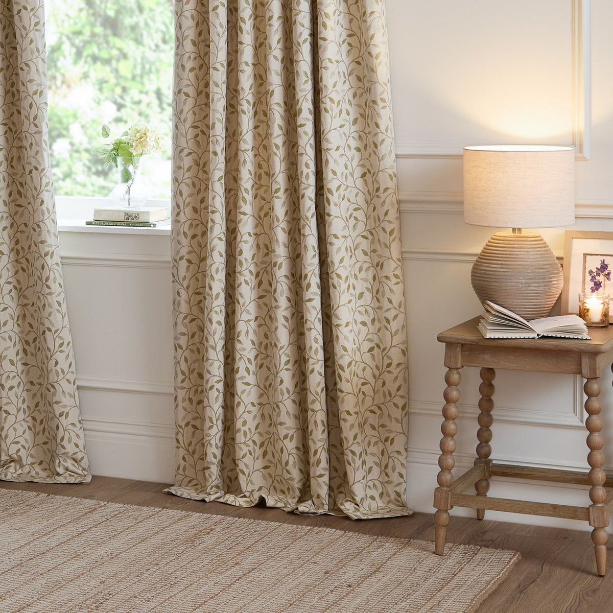 Cervino Jacquard Polyester Pencil Pleat Curtains in Lichen, cream fabric with woven green leaf trail pattern, shown pooling by a living room window beside a wooden side table and lamp.