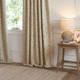 Cervino Jacquard Polyester Pencil Pleat Curtains in Lichen, cream fabric with woven green leaf trail pattern, shown pooling by a living room window beside a wooden side table and lamp.