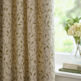 Cervino pencil pleat curtains in Lichen, cream jacquard fabric with woven green leaf and stem pattern, shown pleated beside a window with a vase of flowers on the sill.