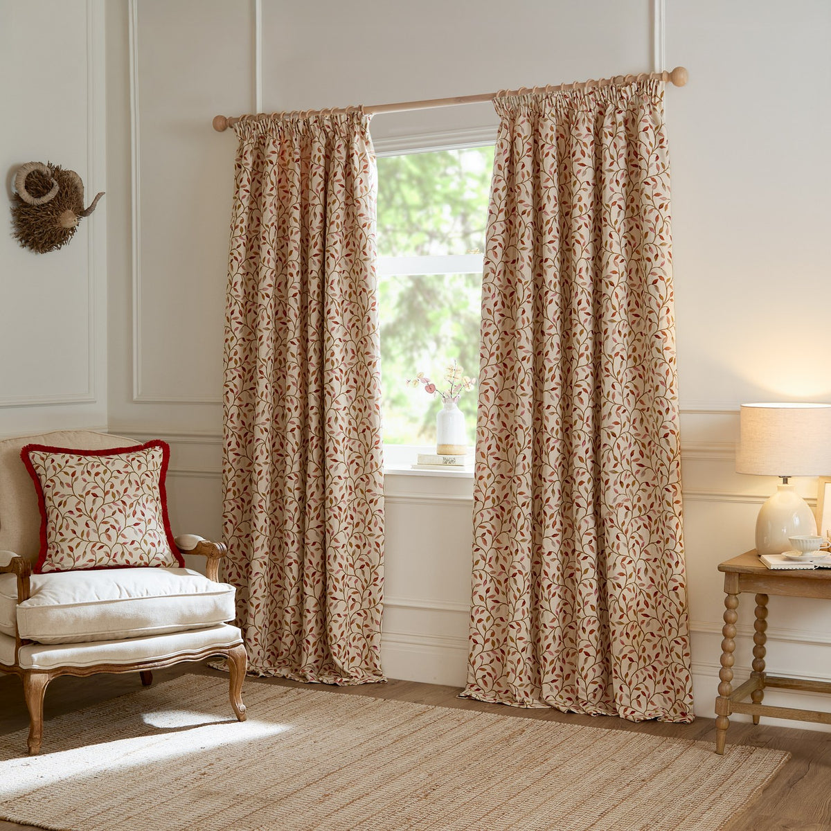 Cervino pencil pleat curtains in cream jacquard fabric with woven leaf and stem pattern in rust, pink and green, shown pleated at a window beside an armchair with matching cushion.
