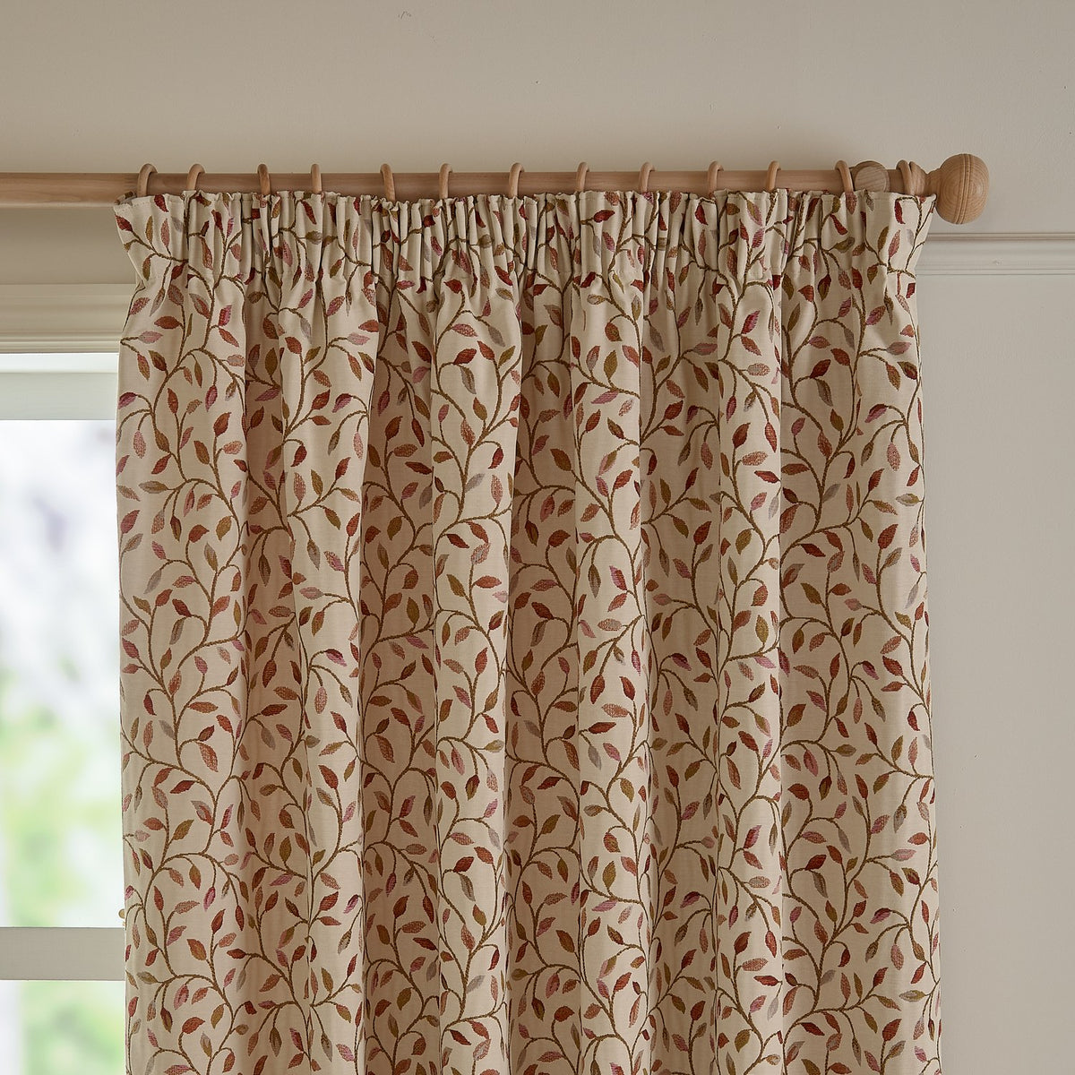 Cervino pencil pleat curtains in cream jacquard fabric with woven leaf and stem pattern in rust, terracotta, pink and green, shown pleated on a wooden pole by a window.