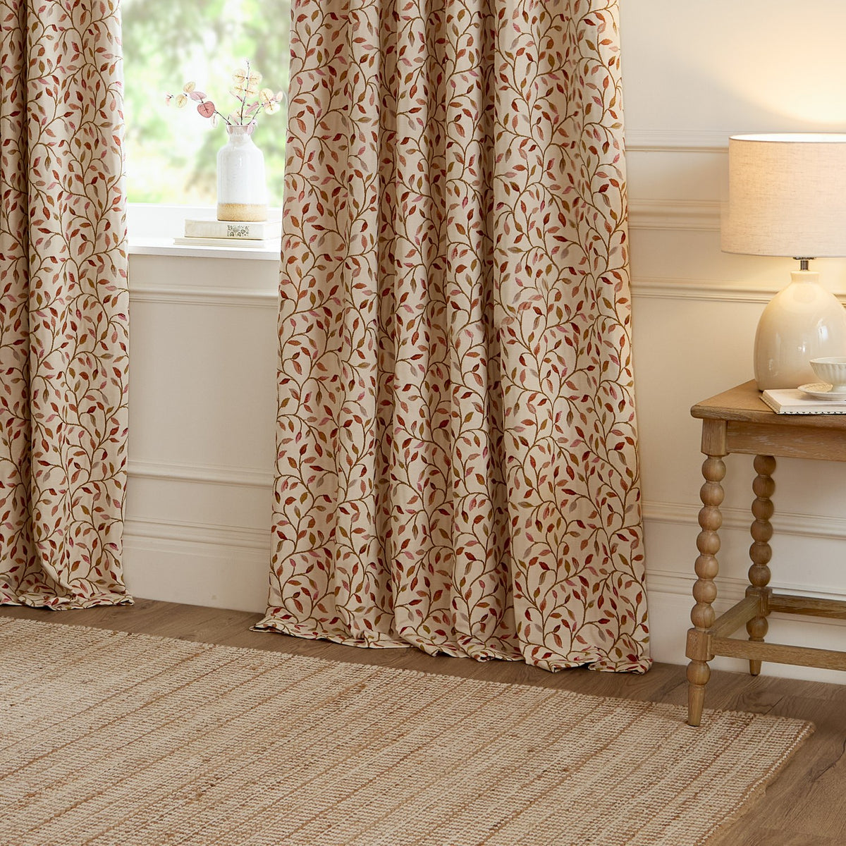 Cervino jacquard pencil pleat curtains in Rednut, cream fabric with woven leaf and stem design in rust, terracotta, pink and green tones, hung at a living room window beside a wooden side table.