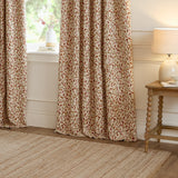 Cervino jacquard pencil pleat curtains in Rednut, cream fabric with woven leaf and stem design in rust, terracotta, pink and green tones, hung at a living room window beside a wooden side table.