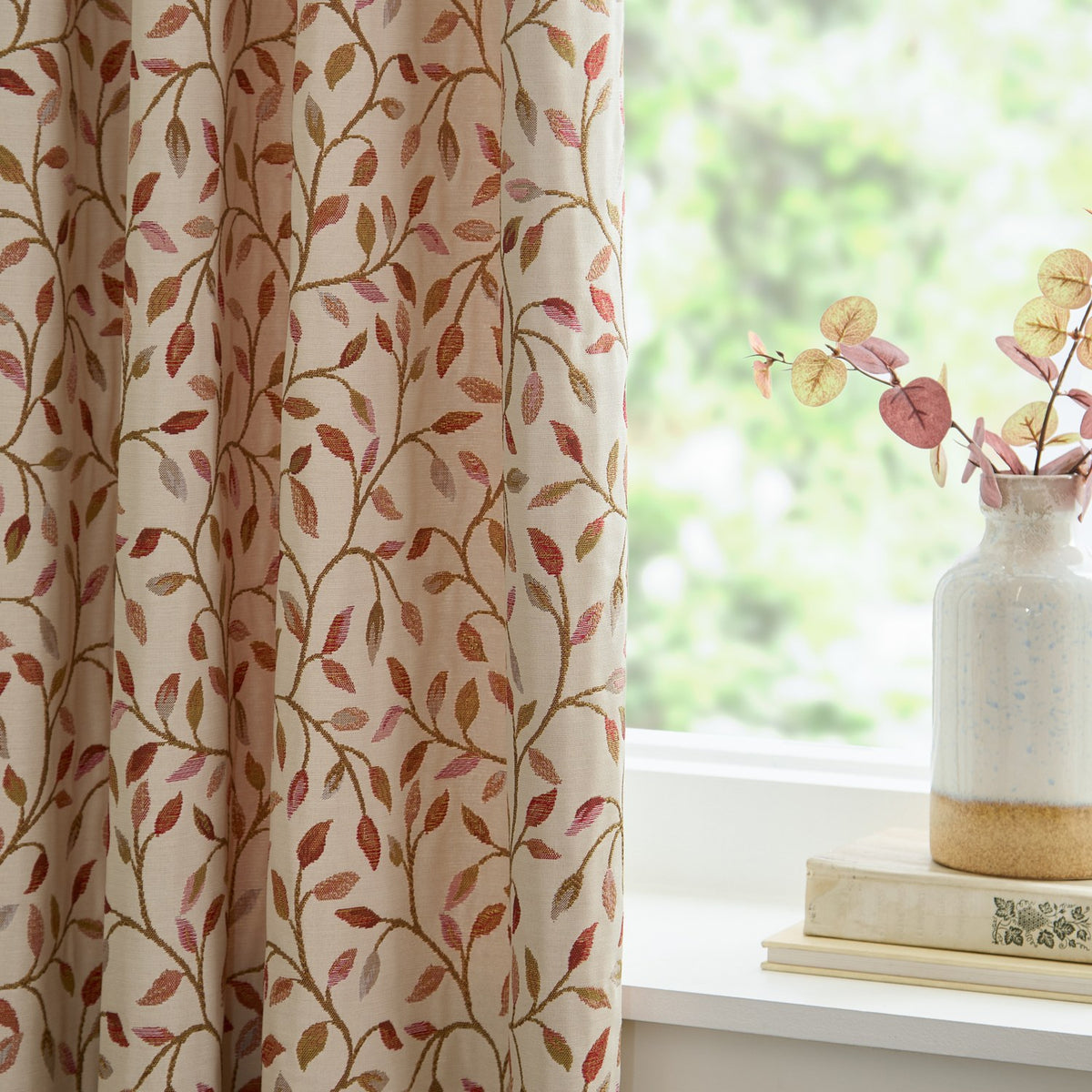 Cervino pencil pleat curtain in cream polyester with jacquard leaf pattern in rust, terracotta, pink and green, shown beside a window with a vase and books on the sill.