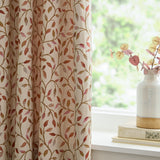 Cervino pencil pleat curtain in cream polyester with jacquard leaf pattern in rust, terracotta, pink and green, shown beside a window with a vase and books on the sill.