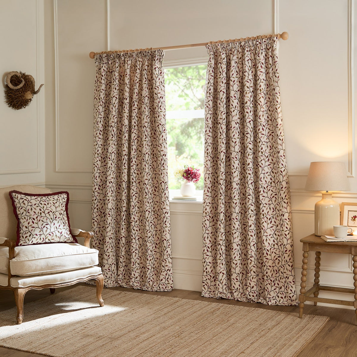 Cervino Jacquard Cotton Blend Cushion in Damson on an armchair, showing a cream ground with leafy trailing pattern in wine, pink and green tones, piped edge, in a living room setting.