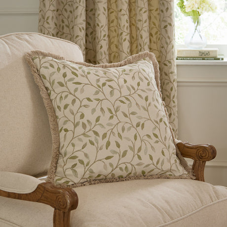 Cervino Jacquard Cotton Blend Cushion in Lichen, a square cream cushion with a woven green leaf pattern and pale sandy fringe trim, shown on a beige armchair.