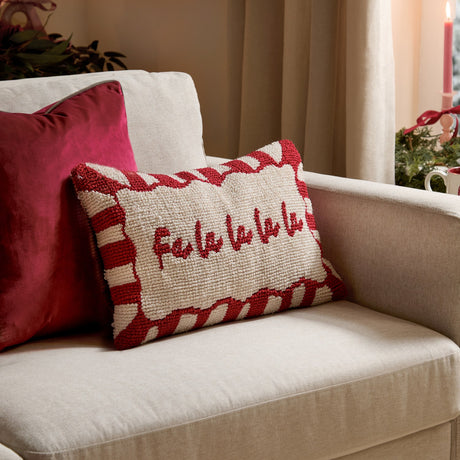 Candy Fa La La cushion with hand-tufted cream face, red "Fa la la la la" script, and red-white candy-stripe border, shown on a beige armchair beside a red velvet cushion.