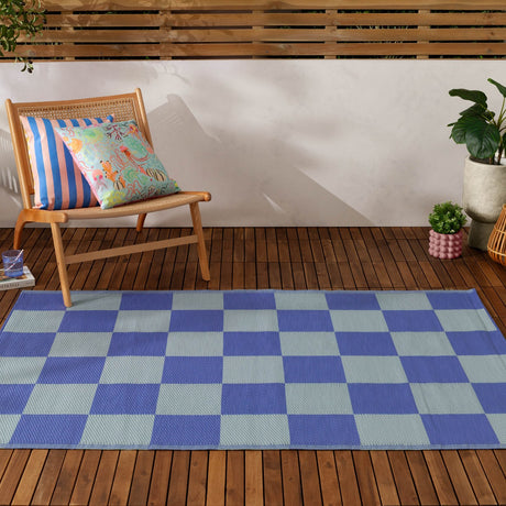 Checkerboard Jacquard Check Rug in Blue, a two-tone periwinkle and duck-egg blue outdoor rug shown on a wooden deck beside a rattan chair with striped cushions.