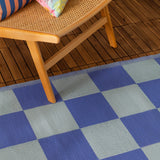 Checkerboard Jacquard Check Rug in blue, woven with periwinkle and duck-egg blue squares, shown on a wooden deck beneath a rattan chair.