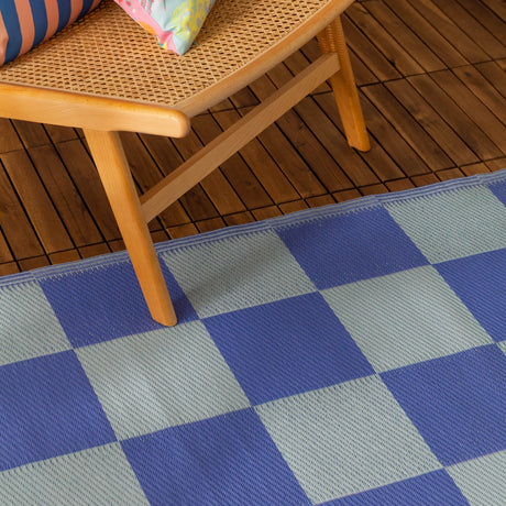 Checkerboard Jacquard Check Rug in blue, woven with periwinkle and duck-egg blue squares, shown on a wooden deck beneath a rattan chair.