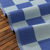 Checkerboard Jacquard Check Rug in Blue, partly rolled to show woven texture, alternating periwinkle and duck-egg blue squares, on a wooden deck.