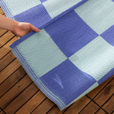 Checkerboard Jacquard Check Rug in Blue, showing woven periwinkle and duck-egg blue squares as a hand lifts the corner on a wooden outdoor deck.