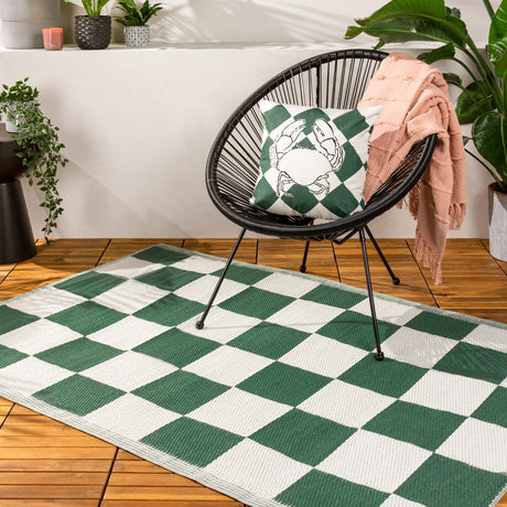 Checkerboard Jacquard Check Rug in green and off-white with striped border, laid on wooden decking beneath a black rattan chair with crab-print cushion.