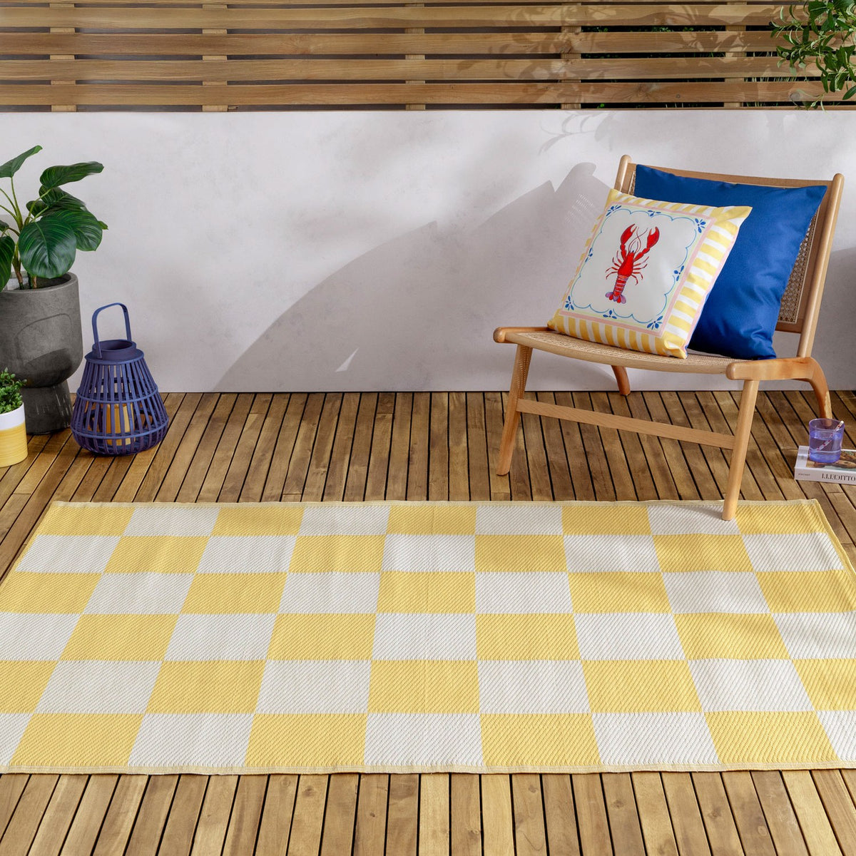 Checkerboard Jacquard Check Rug in yellow and off-white squares, laid on a wooden deck beside a rattan chair with patterned cushions and a plant.