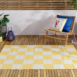 Checkerboard Jacquard Check Rug in yellow and off-white squares, laid on a wooden deck beside a rattan chair with patterned cushions and a plant.