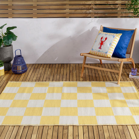 Checkerboard Jacquard Check Rug in yellow and off-white squares, laid on a wooden deck beside a rattan chair with patterned cushions and a plant.
