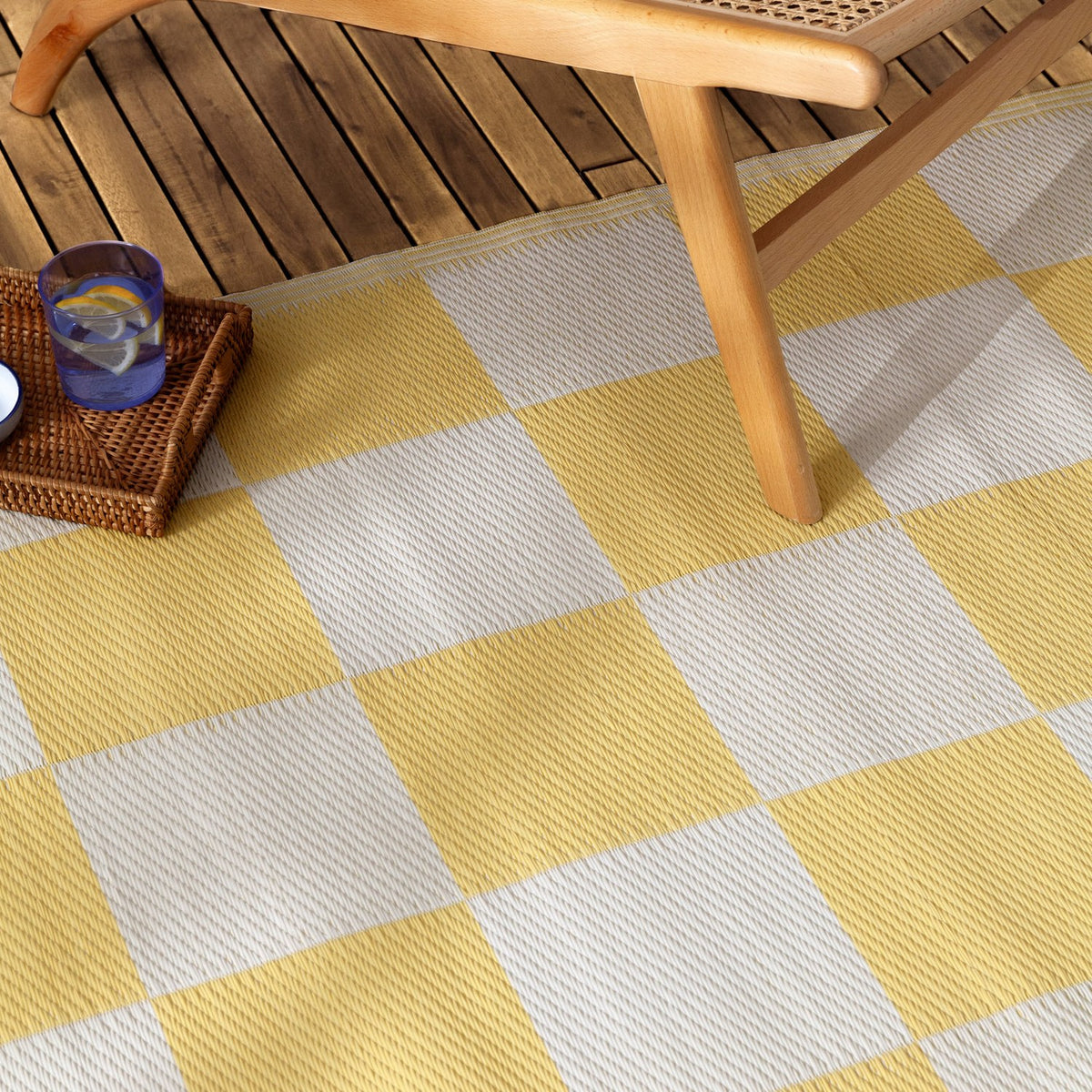 Checkerboard Jacquard Check Rug in yellow and off-white squares, shown on a wooden deck beneath a wooden chair with a rattan tray and glass nearby.