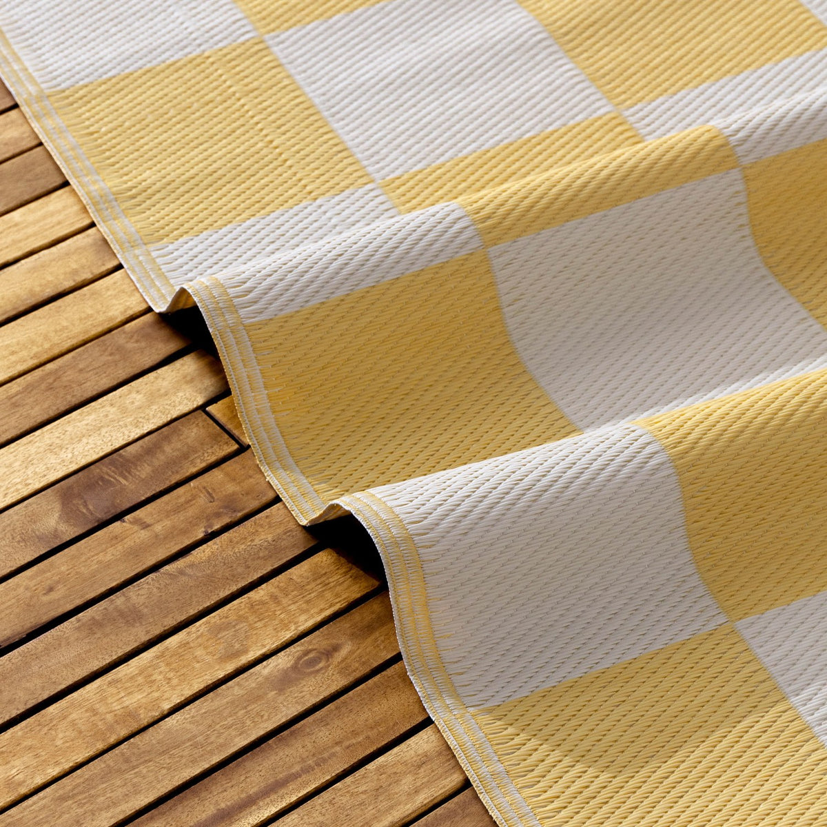 Checkerboard Jacquard Check Rug in yellow and off-white squares, partly rolled to show woven polypropylene texture, laid on a wooden deck.
