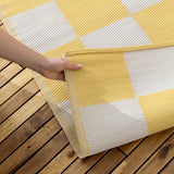 Checkerboard Jacquard Check Rug in Yellow with a bold yellow and off-white large-scale check pattern, shown outdoors on a wooden deck as a hand lifts a corner to reveal the stitched edge.
