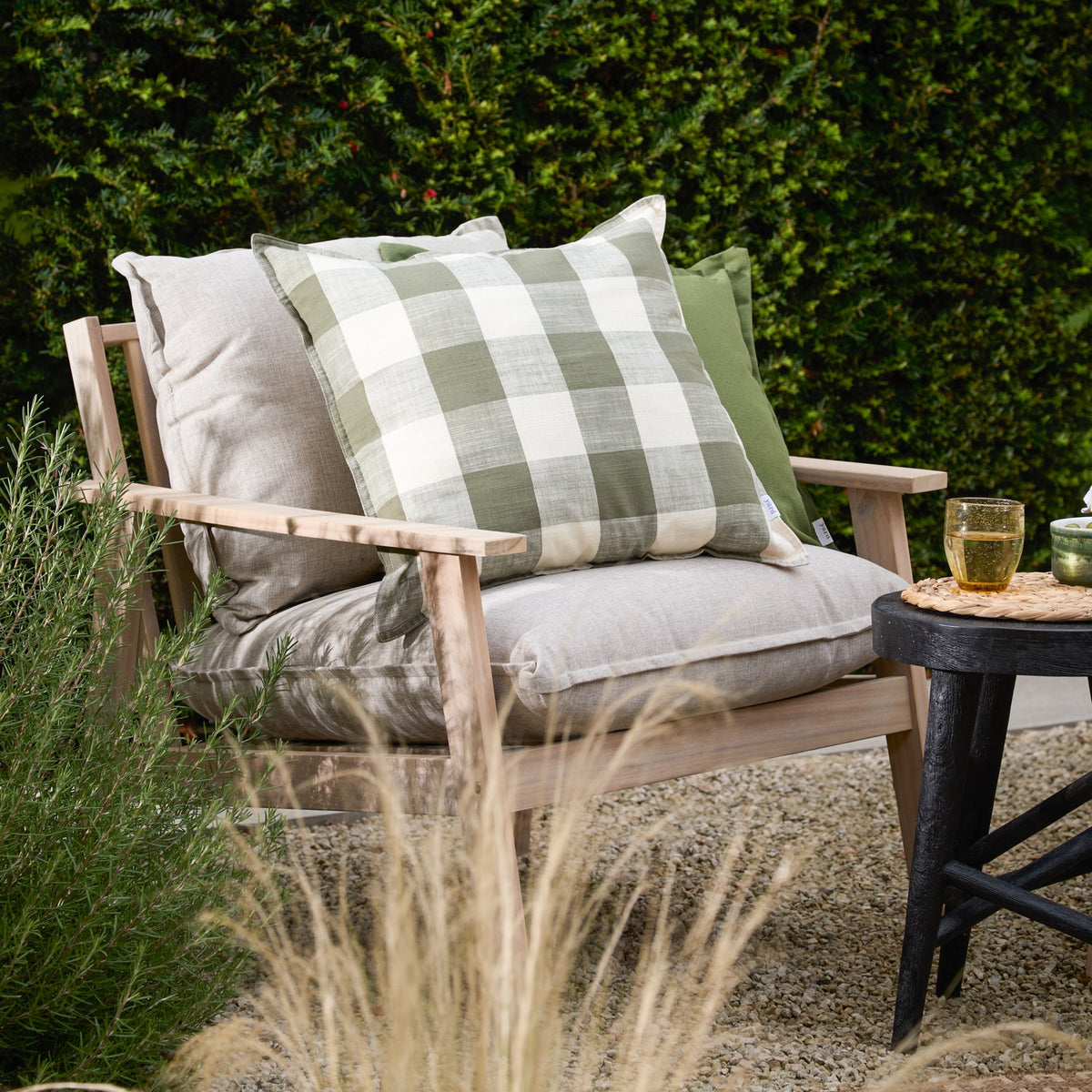 Chesil Check Printed Cotton Cushion in moss green and off-white buffalo check, styled on a wooden outdoor sofa with neutral cushions against a hedge backdrop.