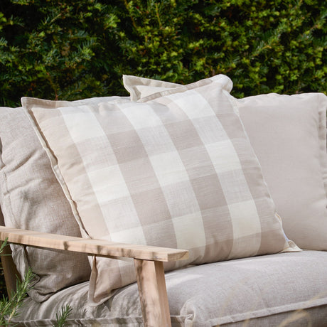 Chesil Check Printed Cotton Cushion in natural beige and off-white, flat oxford border, shown on a neutral sofa in an outdoor garden setting.