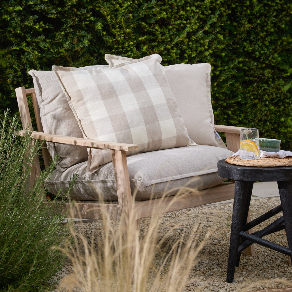 Chesil Check Printed Cotton Cushion in Natural with beige and off-white check pattern and oxford border, shown on a wooden garden bench among plain cream cushions.