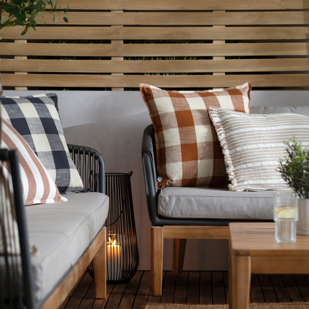 Chesil Check Printed Cotton Cushion in navy and cream buffalo check, styled on an outdoor sofa alongside striped and rust check cushions in a garden setting.