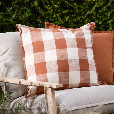 Chesil Check Printed Cotton Cushion in pecan brown and off-white gingham with flat edge trim, styled on a linen outdoor sofa beside a plain terracotta cushion.