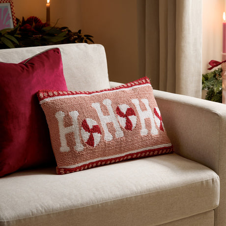 Candy Ho Ho Ho rectangular cushion in blush pink with tufted white "Ho Ho" lettering and red peppermint swirls, red banded borders, shown on a beige sofa with a red cushion.
